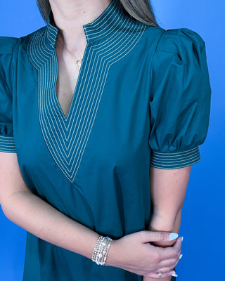 A woman wearing a teal green blouse with a high mock neck, puffed short sleeves, and white chevron stripe detailing on the collar and cuffs, posed against a solid blue background.