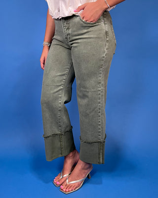 A person wearing washed olive green cropped jeans with rolled cuffs and frayed hems, paired with a white top