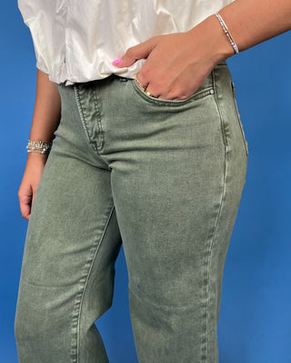 A person wearing faded sage green denim jeans, shown from the waist down against a solid blue background. 