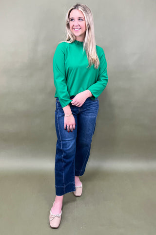Woman wearing a green long-sleeve top and blue jeans against a plain background