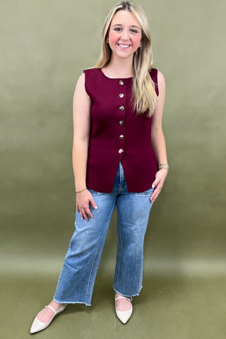 Woman wearing a burgundy sleeveless button-up top and blue jeans against a green background