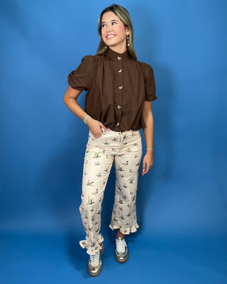 A woman modeling a brown short-sleeve button-up blouse with puff sleeves and cream pants featuring a small bird print, posed against a solid blue studio backdrop.