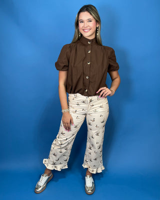 A woman modeling a brown short-sleeve blouse with puff sleeves and decorative gold buttons, paired with beige patterned pant