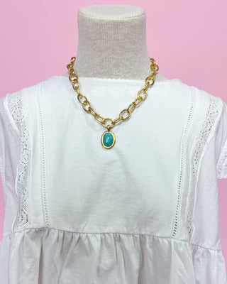 Gold chain necklace with a blue pendant on a white dress against a pink background
