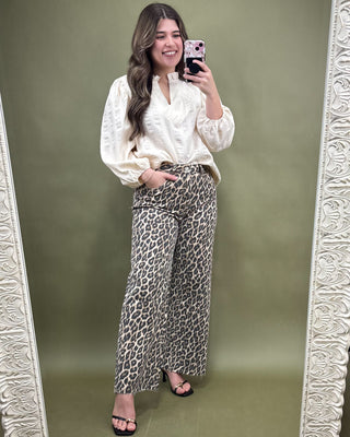 Woman taking a mirror selfie wearing a white blouse and leopard print pants on a green background.