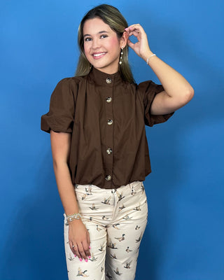 A woman modeling a brown short-sleeve blouse with puff sleeves and decorative gold buttons, paired with beige patterned pant