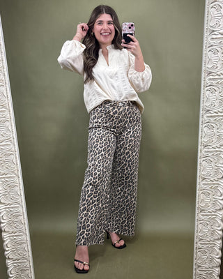 Woman taking a mirror selfie wearing a white blouse and leopard print pants against a green background.