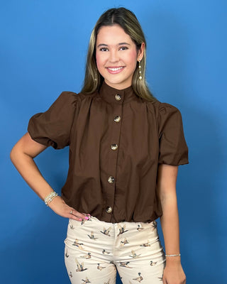 A woman modeling a brown short-sleeve blouse with puff sleeves and decorative gold buttons, paired with beige patterned pant