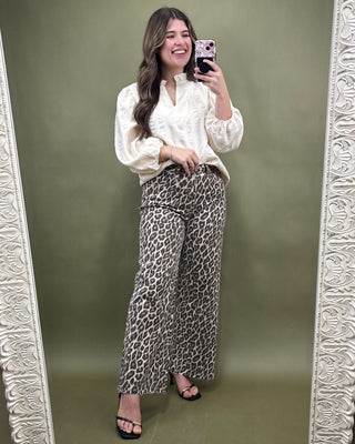 Woman taking a mirror selfie wearing a white blouse and leopard print pants against a green background.