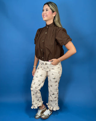A woman modeling a brown short-sleeve button-up blouse with puff sleeves and cream pants featuring a black bird print, posed against a solid blue studio backdrop.