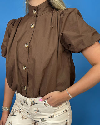 A woman wearing a brown short-sleeve blouse with puff sleeves and a high mock neck, paired with white duck-print shorts, posed against a solid blue background.
