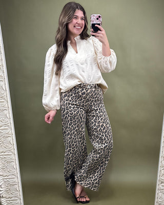 Woman taking a mirror selfie wearing a white blouse and leopard print pants on a green background