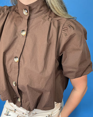 A close-up of a person wearing a brown short-sleeve blouse with a high mock neck and decorative buttons, paired with a light patterned skirt, set against a solid blue background.