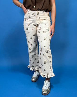 A person wearing cream wide-leg cropped pants with a brown ruffled top and metallic silver sneakers, standing against a solid blue studio background.