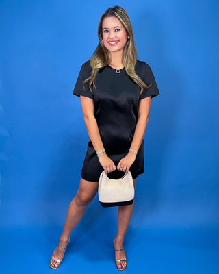 A woman modeling a black satin short-sleeve mini dress with a small cream handbag and metallic sandals, standing against a solid blue studio backdrop.