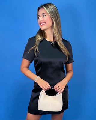 A woman modeling a black satin short-sleeve mini dress with a small white handbag, set against a solid blue studio background.