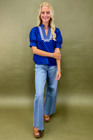 Woman wearing a blue top with embroidery and jeans against a green background