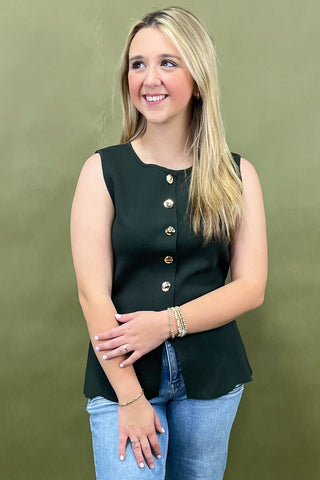 Woman wearing a black sleeveless top with gold buttons against a green background