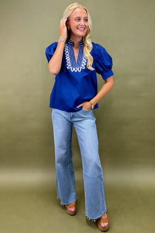 Woman wearing a blue blouse with white embroidery and light blue jeans on a green background