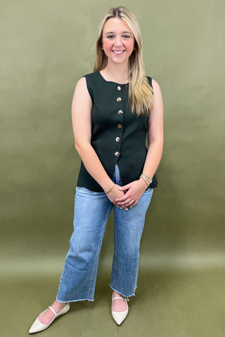 Woman wearing a black sleeveless top with gold buttons and blue jeans against a green background