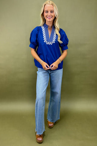 Woman wearing a blue blouse with white embroidery and light blue jeans on a green background