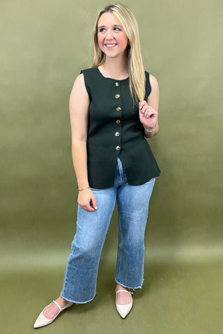 Woman wearing a black sleeveless top with buttons and blue jeans against a green background
