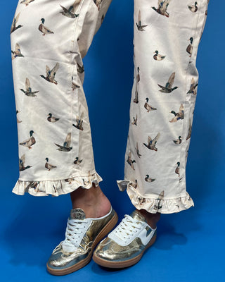 A person wearing cream-colored pants with a brown and green duck print, paired with metallic gold and white sneakers