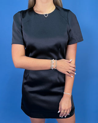 A woman wearing a short-sleeve black satin dress, standing against a solid blue background.