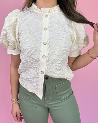 Person wearing a white textured blouse with short sleeves and green pants on a pink background