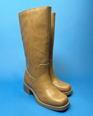 A pair of knee-high taupe faux-leather boots with a low block heel, shown against a solid blue background.