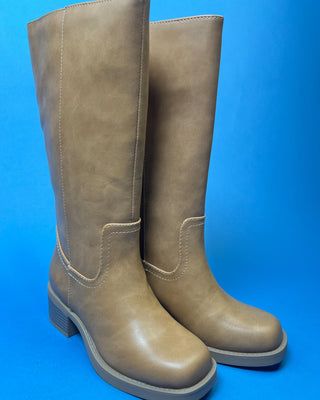 A pair of knee-high taupe faux-leather boots with a low block heel, shown against a solid blue background.