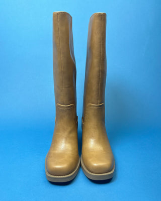 A pair of knee-high taupe faux-leather boots with a low block heel, shown against a solid blue background.