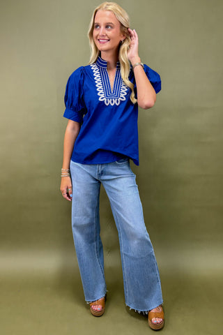 Woman wearing a blue top with white embroidery and jeans on a green background