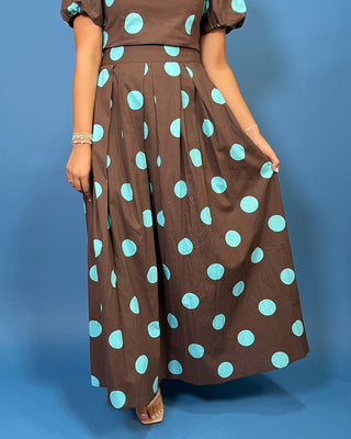 A woman wearing a brown maxi skirt with large light blue polka dots, paired with a matching brown top, standing against a solid blue studio background.
