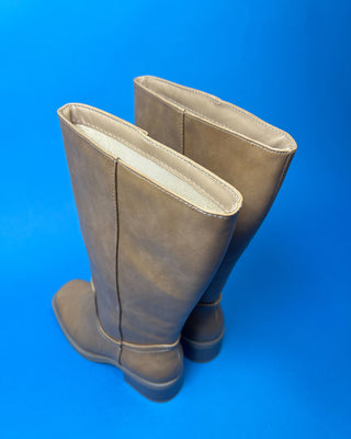 A pair of knee-high taupe faux-leather boots with a low block heel, shown against a solid blue background.