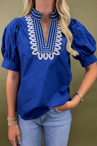 Woman wearing a blue blouse with white embroidery and light blue jeans against a green background