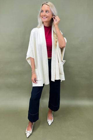 Woman wearing a white cardigan, red top, and dark pants against a green background