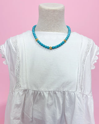 Turquoise beaded necklace on a mannequin with a white dress against a pink background