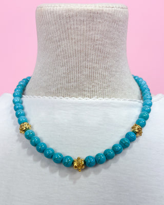 Turquoise beaded necklace with gold accents on a mannequin bust against a pink background