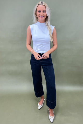 Woman wearing a white sleeveless top and dark blue jeans against a plain background