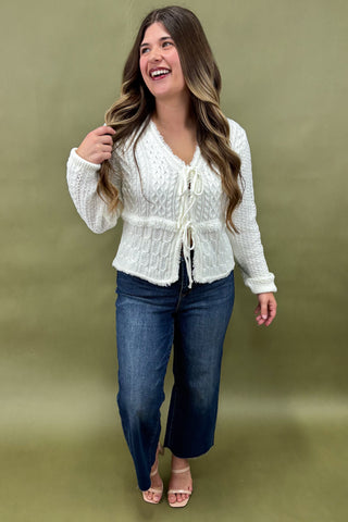 Woman wearing a white knitted cardigan and blue jeans against a green background