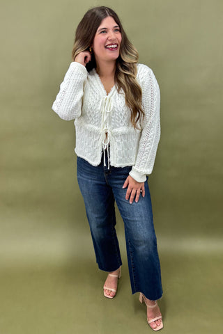 Woman wearing a white knitted cardigan and blue jeans against a green background