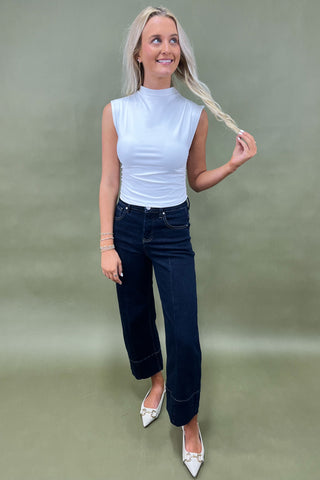 Woman wearing a white sleeveless top and dark blue jeans against a green background