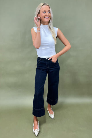 Woman wearing a white sleeveless top and dark blue jeans against a green background
