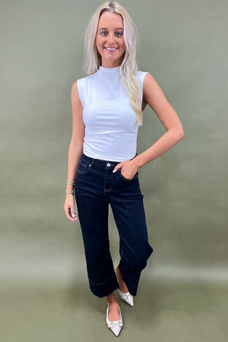 Woman wearing a white sleeveless top and dark jeans against a plain background