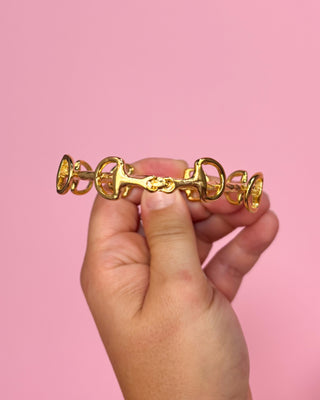 Gold horse bit held between fingers against a pink background