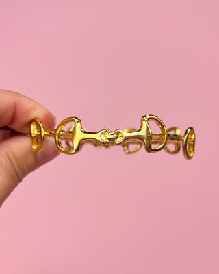 Gold bracelet held by a hand against a pink background