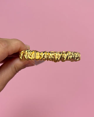 Gold bangle held by a hand against a pink background