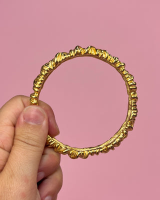 Gold bracelet held by a hand against a pink background