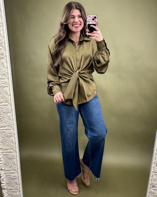 Woman taking a mirror selfie wearing an olive green shirt and blue jeans on a green background